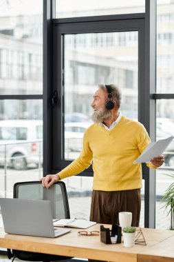 Handsome mature man with headphones smiles while holding documents in a bright office. clipart