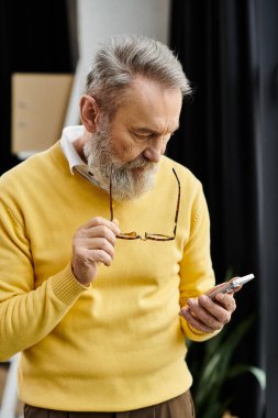 Handsome senior man wearing a yellow sweater looks at his smartphone with concentration. clipart