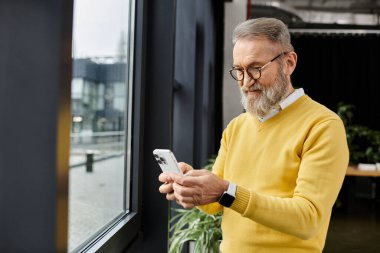 Sarı kazaklı yakışıklı bir adam pencerenin yanındaki akıllı telefonuna odaklanıyor..
