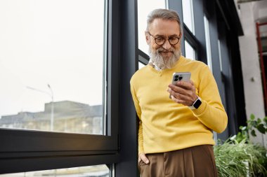 Sarı kazak giyen olgun, yakışıklı bir adam büyük bir pencereden akıllı telefonuna bakarken gülümsüyor..