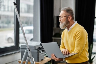 Olgun ve yakışıklı bir adam bir sehpanın yanında dururken dizüstü bilgisayarına odaklanır..