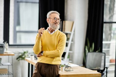 Bearded man in glasses stands thoughtfully in a stylish office, dressed casually. clipart