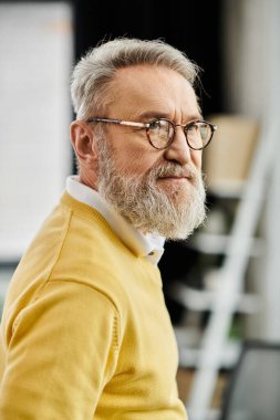 Sakallı ve şık gözlükleri olan olgun bir adam parlak bir ofis ortamında düşünür..