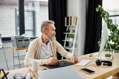 Mature man with a beard engaged in work at a stylish desk in a well lit office space clipart