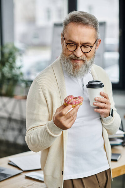 A stylish senior man holds a donut and coffee while taking a moment to relax at work.