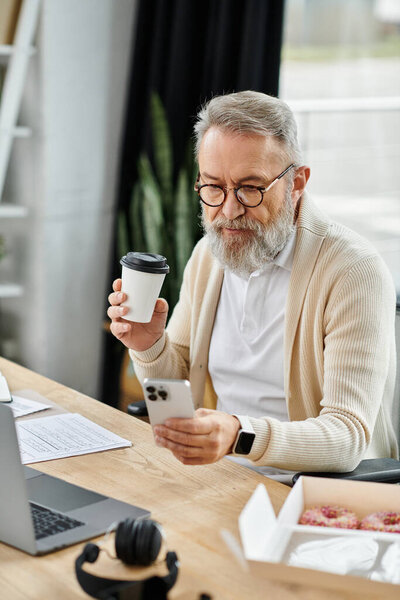 Красивый пожилой мужчина пьет кофе и проверяет свой смартфон в стильном офисе.