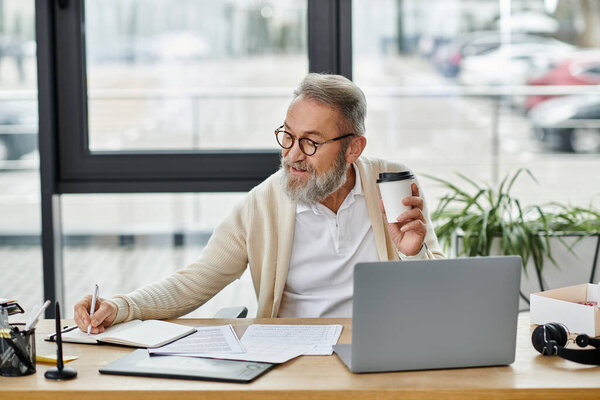 A mature handsome man with glasses writes notes and sips coffee at a modern desk.
