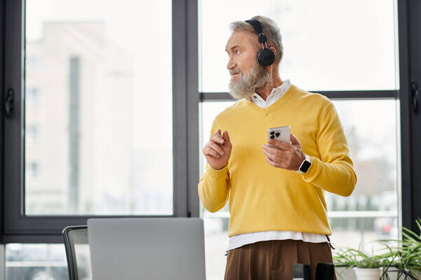 Senior man wearing headphones engages with his smartphone in a bright office environment.