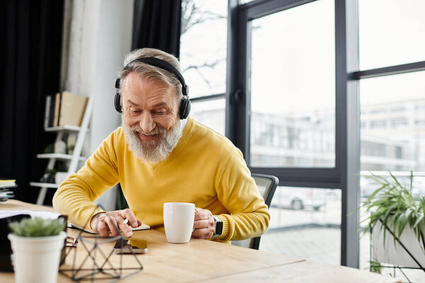 Handsome senior man smiles while sipping coffee and using a smartphone at his desk.