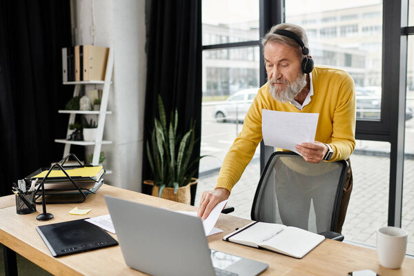 A handsome man at a desk examines papers intently while wearing headphones.