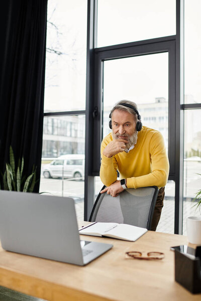 A mature man in a yellow sweater focuses on a laptop during a video call at his office.