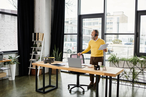 A mature handsome man stands in a bright office, reviewing documents and enjoying the view outside.