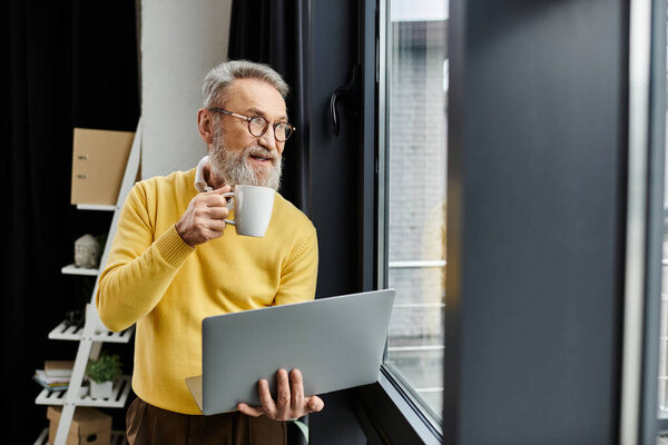 Mature man sips coffee and works on a laptop, gazing out the window in a cozy room.