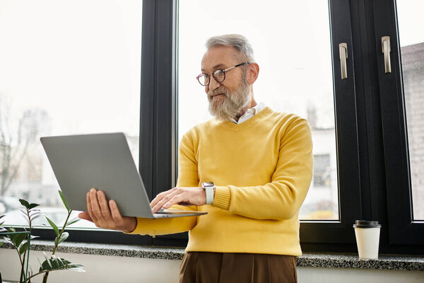 A senior man in a yellow sweater collaborates on a laptop while standing by a window.