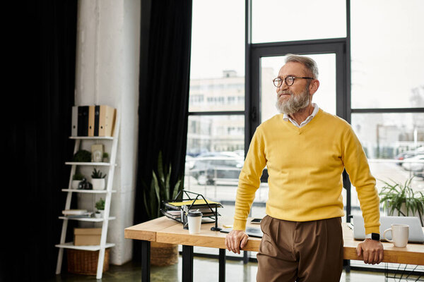 A handsome man in a yellow sweater stands confidently in a modern office, looking outside.