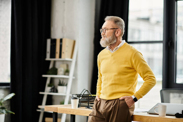 Взрослый красивый мужчина в жёлтом свитере стоит у стола в современном офисе.