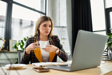 Kareli ceketli çekici bir kadın kahvesini yudumlarken dizüstü bilgisayarıyla meşgul oluyor..