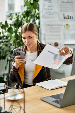 İşle meşgul olan genç bir kadın, masasındaki dosyaları incelerken telefonundaki verileri inceliyor..