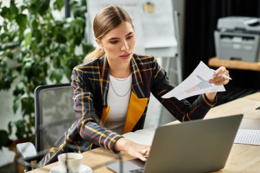 Attractive young woman focuses on her laptop while reviewing documents in a contemporary workspace. clipart