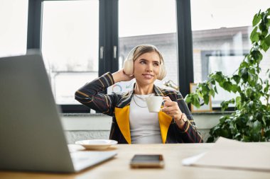 Attractive woman in a checkered blazer relaxes with coffee while managing work tasks. clipart