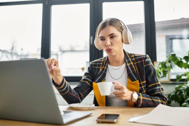 Focused young professional in checkered blazer using laptop and enjoys coffee during virtual meeting. clipart