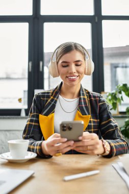 Dişli ceketli, kendine güvenen bir kadın modern ofis ortamında akıllı telefonunu kullanırken gülümser..