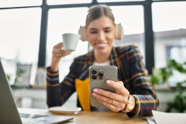 Stylish young woman in a checkered blazer engages with her smartphone while sipping coffee. clipart