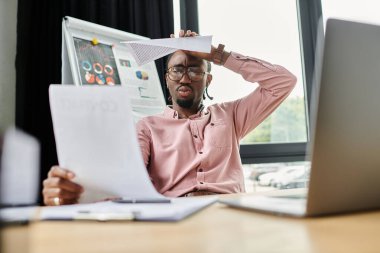 A young African American man reviews documents with a thoughtful expression in a modern workspace. clipart