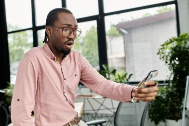 Genç Afro-Amerikalı bir adam, canlı bir ofiste telefonunu kontrol ederken şaşkınlıkla tepki veriyor..