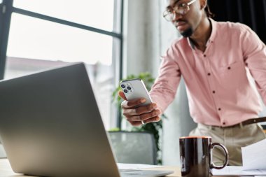 A young african american man uses his smartphone while focused on his laptop in a bright workspace. clipart