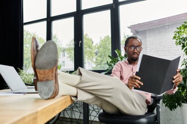 A young african american man relaxes at a stylish desk, reviewing documents in a coworking space. clipart