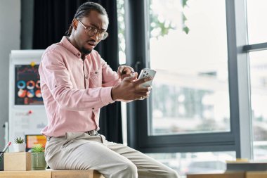 Genç Afrikalı Amerikalı adam modern bir çalışma alanında otururken akıllı telefonuna odaklanmış..