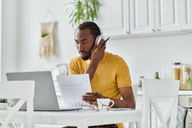 Focused young man reviews documents while working remotely in a stylish kitchen space. clipart