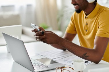 A young african american man in a yellow shirt is working remotely on his phone and laptop. clipart