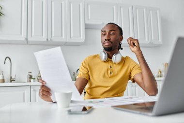 Focused young man reviews documents in a stylish kitchen, immersed in his remote work. clipart