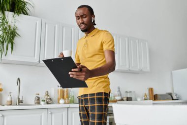 A young man reviews his clipboard while holding coffee in his stylish kitchen. clipart