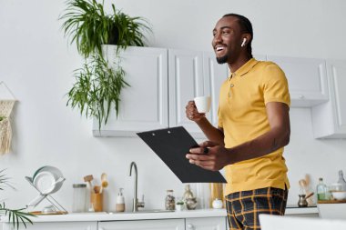 Gülümseyen Afrikalı Amerikalı genç adam, elinde bir bardak ve pano tutarken evde uzaktan çalışmalar yapıyor..