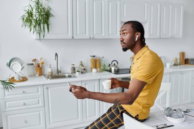 Modern bir mutfakta telefonuyla uğraşırken Airpod 'lu genç bir adam kahvesini yudumluyor..