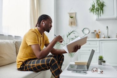 Odaklanmış genç adam rahat bir kanepede oturur, şık bir odada laptopuyla çalışırken notlar alır..