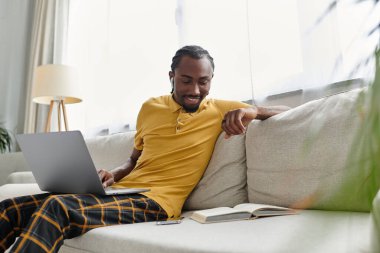 A young African American man focuses on his laptop with a notebook nearby in a bright living space. clipart