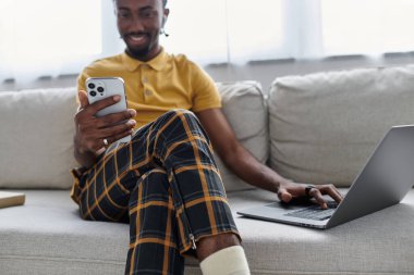 Genç Afrikalı Amerikalı bir adam dizüstü bilgisayarı üzerinde çalışırken kanepede rahatlıyor ve telefonunu kontrol ediyor..