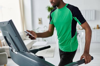 A young man works out on a treadmill in a bright, modern room, using his phone. clipart