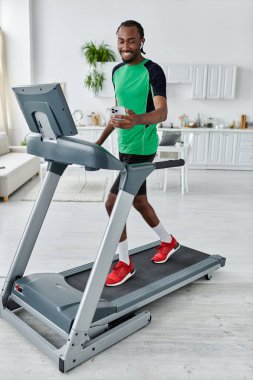 Yeşil spor elbiseli bir adam evdeki koşu bandında yürüyor, telefonunu kullanırken gülümsüyor..