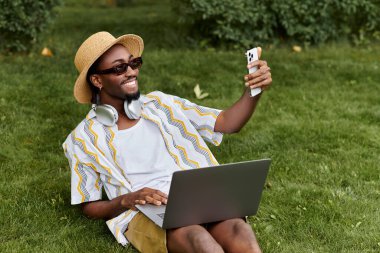 A young african american man relaxes on grass, working on his laptop and taking selfies. clipart