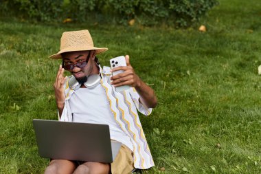 Young man enjoys remote work on his laptop in a vibrant park while using his smartphone. clipart