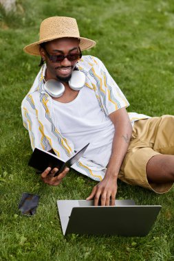 Genç adam, bilgisayarını ve tabletini kullanarak yemyeşil bir parkta uzaktan çalışmayı seviyor..