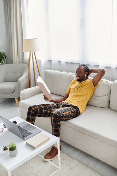 Молодой африканский американец удобно сидит на современном диване, занимается удаленной работой, улыбается.