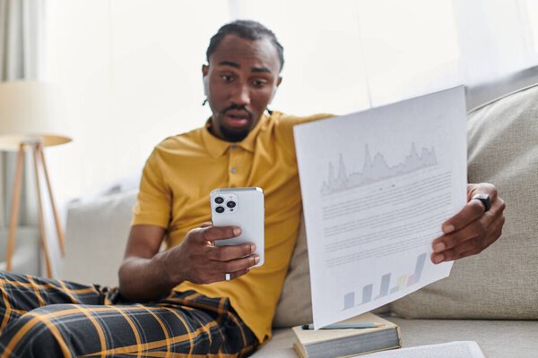 A young african american man reviews data on his smartphone, fully focused while sitting at home.