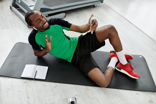 Веселый молодой человек вступает со своим устройством, находясь на гимнастическом коврике, смешивая фитнес и работу плавно.