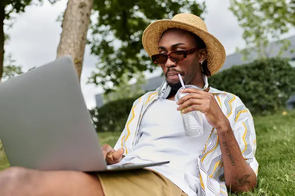 Genç bir Afrikalı Amerikalı, parkta içki içerken bilgisayarında uzaktan çalışmaktan zevk alıyor..